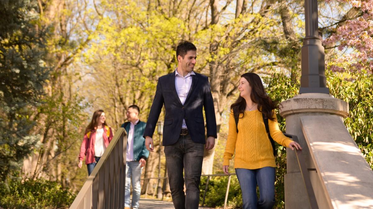 A female in jeans and a gold sweater talks with a male in a blazer as they walk down Hello Walk on the University of Idaho campus.